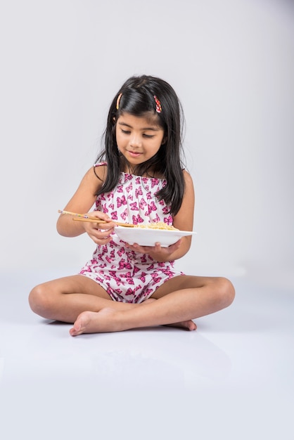 Mignonne petite fille indienne ou asiatique mangeant de délicieuses nouilles chinoises avec une fourchette ou des baguettes, isolée sur fond coloré