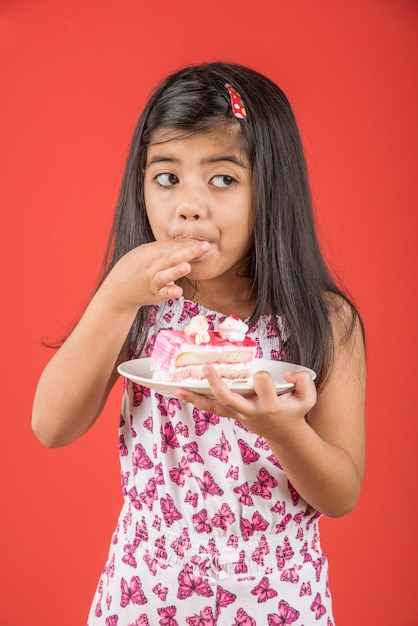 Mignonne petite fille indienne ou asiatique mangeant un morceau de pâtisserie ou de gâteau à la fraise ou au chocolat dans une assiette. Isolé sur fond coloré