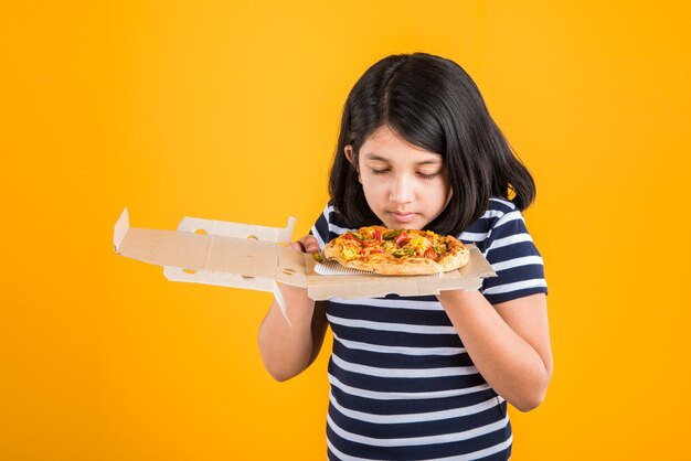 Mignonne petite fille indienne ou asiatique mangeant de savoureux hamburgers, sandwichs ou pizzas dans une assiette ou une boîte. Debout isolé sur fond bleu ou jaune.