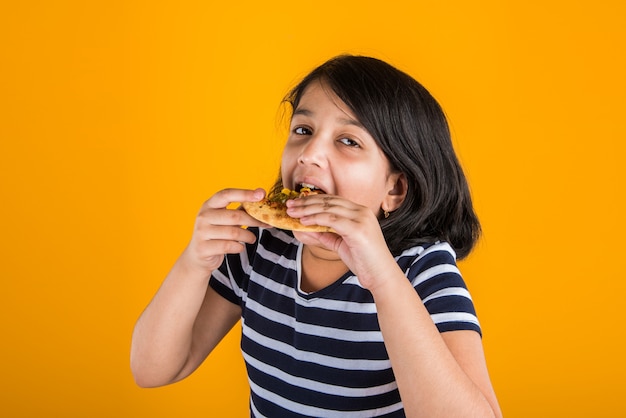 Mignonne petite fille indienne ou asiatique mangeant de savoureux hamburgers, sandwichs ou pizzas dans une assiette ou une boîte. Debout isolé sur fond bleu ou jaune.