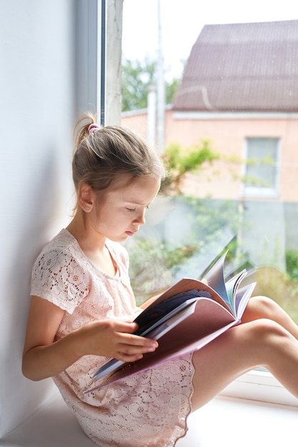 Mignonne petite fille lisant un livre à la maison, au rebord de la fenêtre