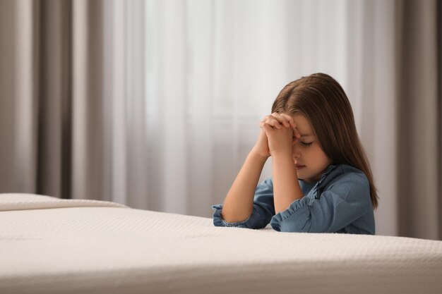 Mignonne petite fille avec les mains jointes disant la prière du coucher dans la chambre Espace pour le texte