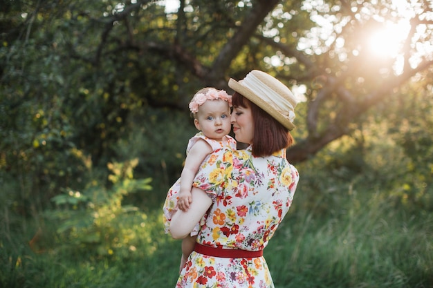 Mignonne petite fille et maman s'embrassent dans le jardin d'été. Vue arrière de la jolie femme mère tenant son bébé sur les mains. La petite fille regarde la caméra. Famille heureuse, maman et fille sur le terrain