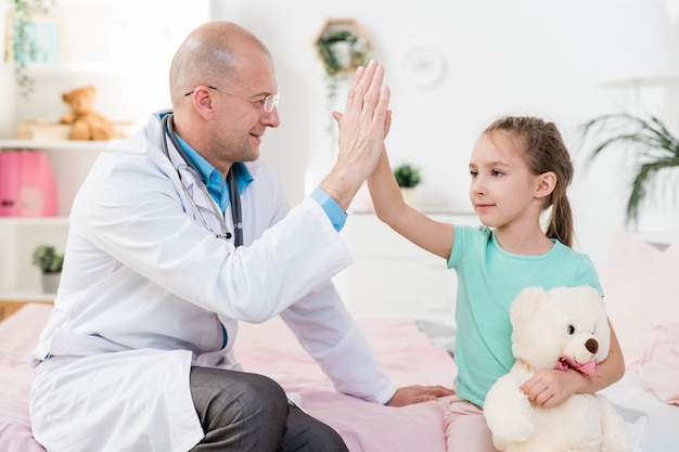 Mignonne petite fille avec ours en peluche et son médecin en blanchon se donnant cinq en position assise à l'hôpital