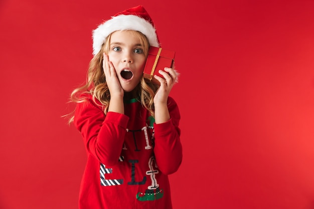 Mignonne petite fille portant un chapeau de Noël debout isolé, tenant la boîte présente