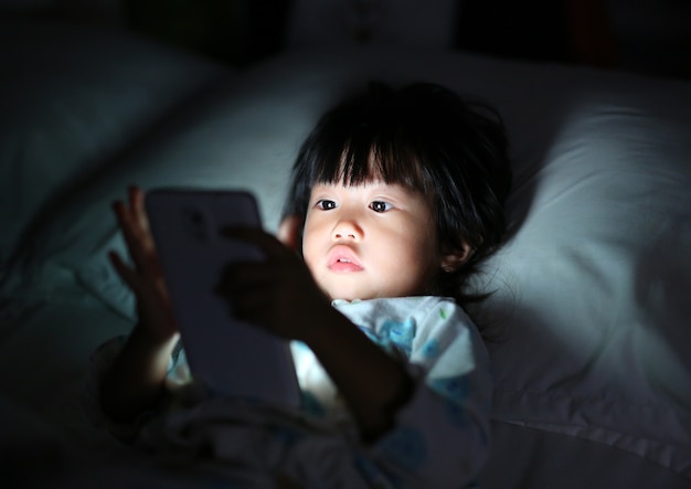 Mignonne petite fille en pyjama jouant smartphone couché sur un lit