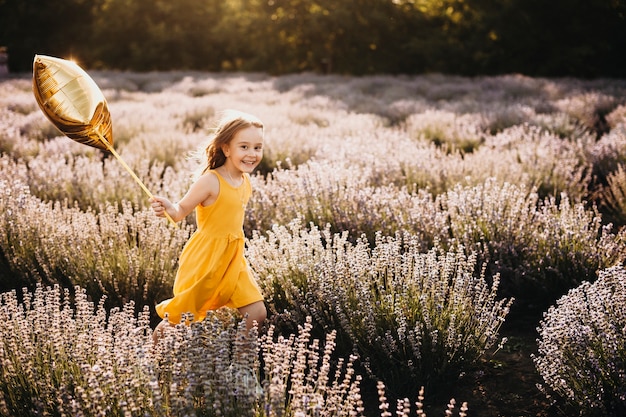 Mignonne petite fille regardant la caméra en souriant tout en courant tenant un ballon dans un champ de fleurs.