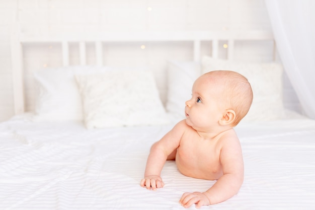 Mignonne petite fille regarde sur le côté dans un berceau sur un lit en coton blanc allongé sur le ventre dans la pépinière