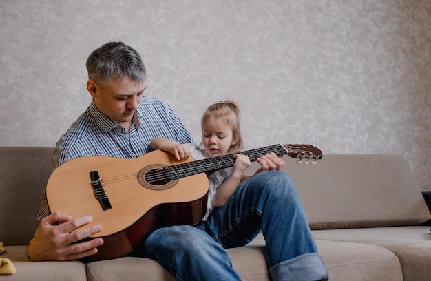 Mignonne petite fille et son beau père jouent de la guitare et sourient assis sur le canapé à la maison. fête des pères. soins et éducation des enfants.