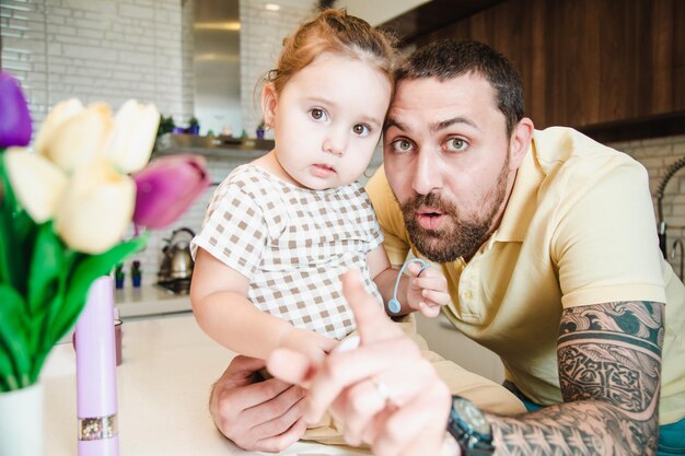 Mignonne petite fille avec son père heureux à la recherche d'un appareil photo étonné dans la cuisine à la maison s'amusant ensemble concept de famille heureuse