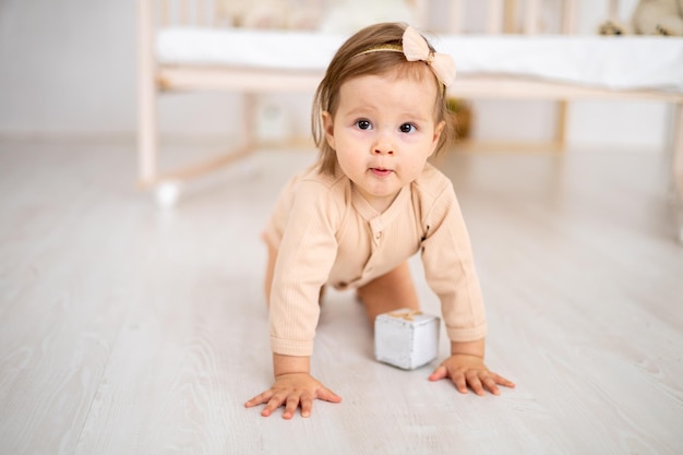 Mignonne petite fille souriante en bonne santé en body beige assise sur le sol près du berceau à la maison dans la pépinière jouant avec des cubes en bois développement des enfants