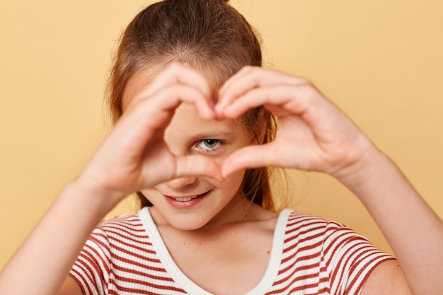 Mignonne souriante optimiste petite fille aux cheveux bruns portant un t-shirt rayé debout isolé sur fond beige montrant la forme de coeur avec les mains devant son visage