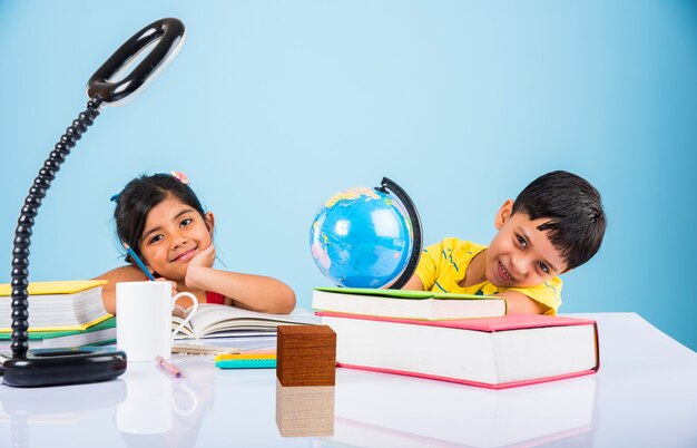 Mignons petits enfants indiens ou asiatiques étudiant sur une table d'étude avec une pile de livres, globe éducatif, isolé sur une couleur bleu clair