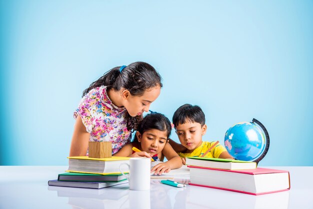Mignons petits enfants indiens ou asiatiques étudiant sur une table d'étude avec une pile de livres, globe éducatif, isolé sur une couleur bleu clair