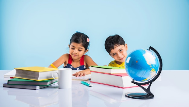 Mignons petits enfants indiens ou asiatiques étudiant sur une table d'étude avec une pile de livres, globe éducatif, isolé sur une couleur bleu clair