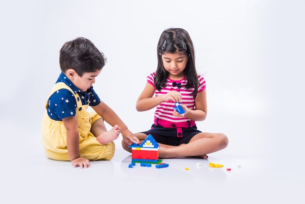 Mignons petits enfants indiens ou asiatiques jouant avec des jouets ou des blocs et s'amusant assis à table ou isolés sur fond blanc