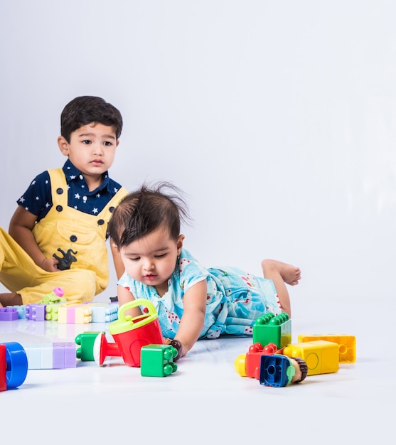 Mignons petits enfants indiens ou asiatiques jouant avec des jouets ou des blocs et s'amusant assis à table ou isolés sur fond blanc