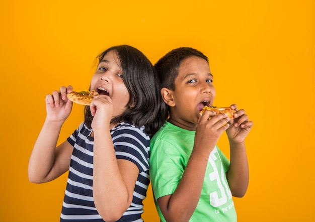 Mignons petits enfants indiens ou asiatiques mangeant de délicieux hamburgers, sandwichs ou pizzas dans une assiette ou une boîte. Debout isolé sur fond bleu ou jaune.