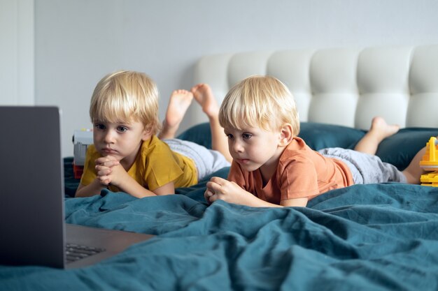 Mignons petits jumeaux de garçons d'enfants caucasiens utilisant un ordinateur portable au lit à la maison. Enfants utilisant la technologie.