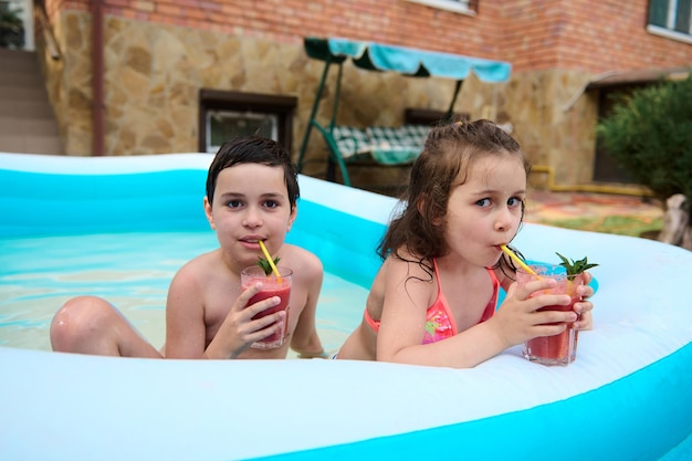 De mignons tout-petits assis dans une piscine d'eau gonflable et buvant du jus de fraise fraîchement pressé à partir d'une paille à boire