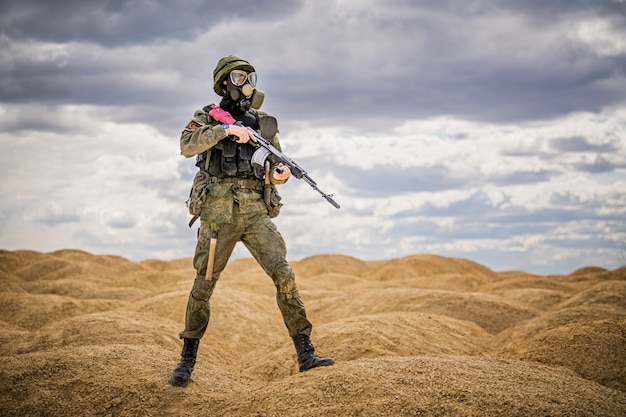 Un militaire dans un masque à gaz avec une arme se dresse au milieu des collines de sable