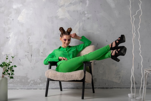 Mince élégante jeune femme sur talons et costume vert est assis sur une chaise sur fond de mur gris en studio