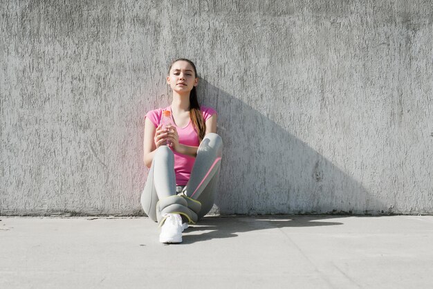 Mince jeune fille fatiguée dans un t-shirt rose assis sur le sol et se reposant après une séance d'entraînement