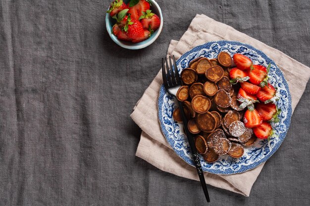 Mini céréales crêpes au chocolat avec des fraises pour le petit-déjeuner sur une vieille nappe textile grise. Petit-déjeuner maison tendance avec de petites crêpes. Vue de dessus