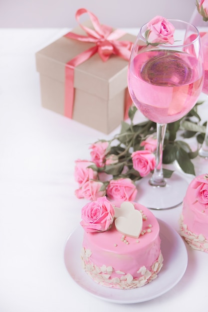 Mini petit gâteau avec glaçage rose, belles roses, tasse de café, coffret cadeau sur le tableau blanc.