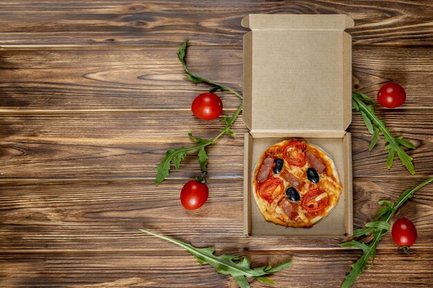 Mini pizza préparée par un enfant avec tomates, olives et jambon sur un fond en bois.