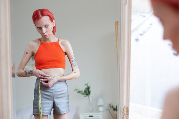 Miroir et tour de taille. Femme rousse anorexique avec des tatouages regardant dans un miroir et mesurant le tour de taille