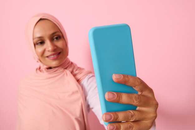 Mise au point douce sur le smartphone en couverture bleue dans les bras tendus d'une femme arabe musulmane portant une tenue islamique religieuse traditionnelle, un hijab rose et des sourires avec un sourire à pleines dents faisant un autoportrait, selfie