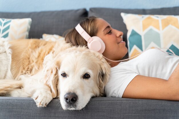 Mise au point sélective sur une femme caucasienne écoutant de la musique avec un chien à la maison. Vue latérale d'une jeune femme avec un casque pris en charge sur son chien labrador dans le canapé. Technologie, personnes et modes de vie des chiens.