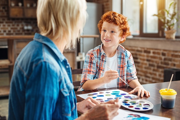 Mise au point sélective sur un garçon excité souriant à sa grand-mère tout en ayant une conversation agréable et en peignant à une table.