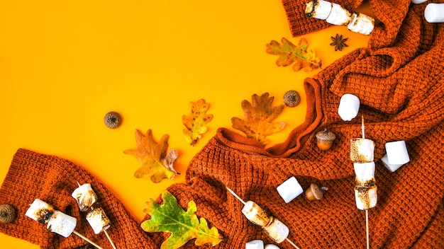 Mise à plat d'automne avec pull orange tricoté, guimauves grillées et feuilles sur fond jaune, vue de dessus. Copiez l'espace pour le texte. Ornement pour Thanksgiving. Cadre d'automne