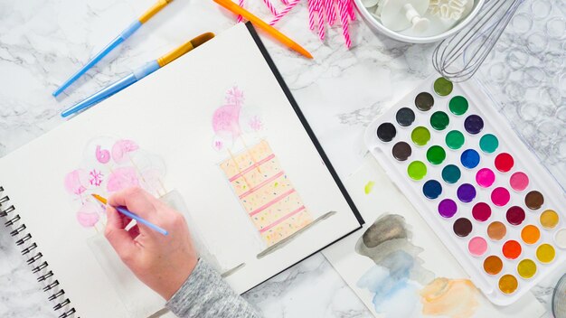 Mise à plat. Baker dessine le dessin d'un gâteau d'anniversaire avec des aquarelles.