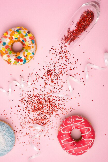 Mise à plat de la célébration. verre de champagne avec des banderoles colorées et de délicieux beignets sur fond rose.