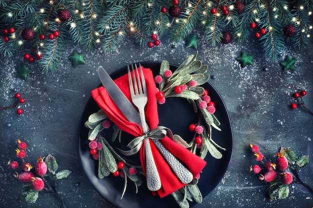 Mise à plat avec des décorations de Noël en vert et rouge avec des baies givrées, des bibelots, des assiettes noires et de la vaisselle