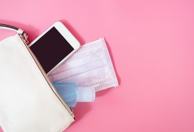 Mise à plat du sac femme en cuir blanc avec smartphone, masque chirurgical et désinfectant au gel d'alcool sur fond rose, vue de dessus avec espace de copie pour le texte. Concept Covid-19 et coronavirus.