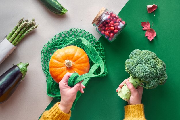 Mise à plat écologique zéro déchet avec les mains tenant le brocoli et le sac de ficelle avec la citrouille orange. Tomber à plat avec des légumes et des mains sur fond de papier kraft, espace de texte.