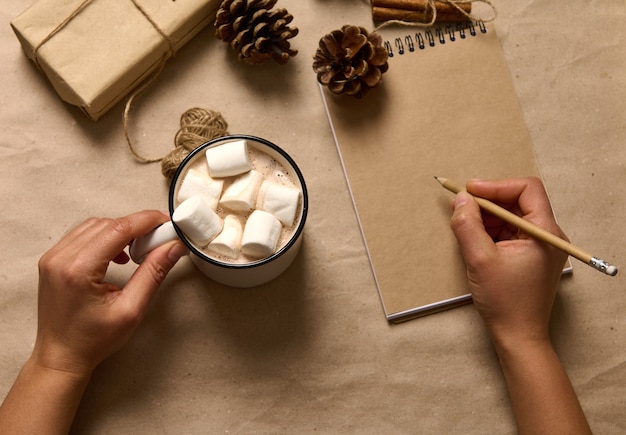 Mise à plat. Des mains humaines tiennent une tasse avec une boisson chaude au cacao et des guimauves et écrivent avec un crayon en bois sur une feuille de cahier vierge sur la surface en carton avec un cadeau de Noël artisanal et des pommes de pin