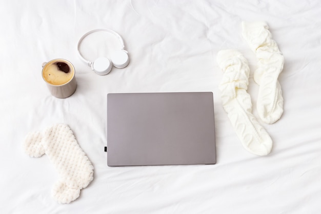 Mise à plat avec ordinateur portable, chaussettes chaudes, tasse de café, écouteurs, masque pour dormir sur le lit dans la chambre