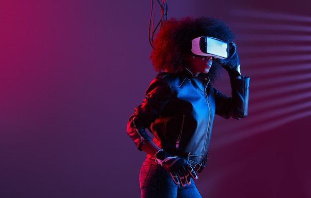 Mod fille aux cheveux noirs bouclés vêtue d'une veste en cuir noir et de gants utilise les lunettes de réalité virtuelle sur sa tête dans le studio sombre avec un néon. Personnage cyberpunk