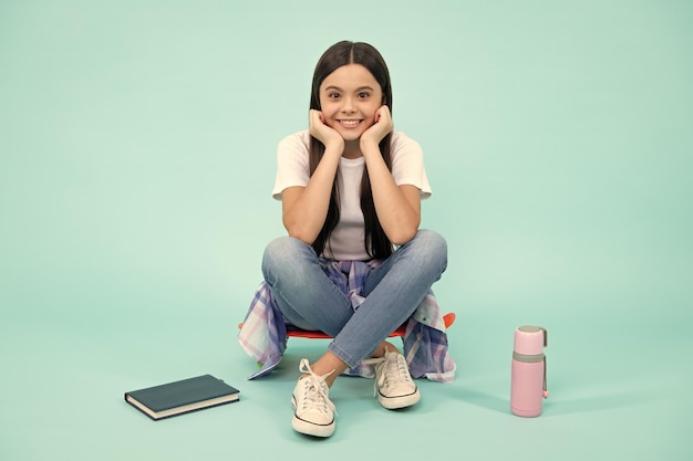 Mode adolescente portant un t-shirt à la mode et un jean s'asseoir sur une planche à roulettes posant sur fond bleu