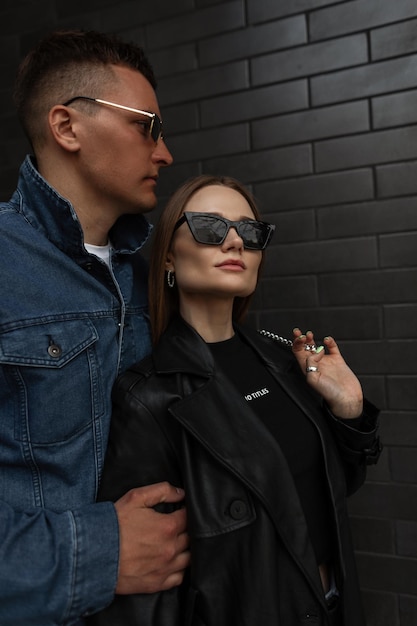 À la mode beau couple femme et homme en tenue de mode avec des lunettes de soleil à la mode en manteau noir et veste en jean se dresse et pose près d'un mur de briques noires dans la rue