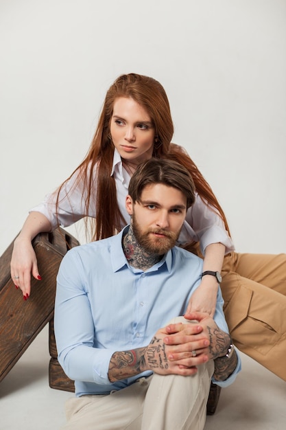 À la mode beau jeune couple en studio sur le sol Beau mec branché hipster avec tatouages et barbe et belle fille rousse élégante assis et posant sur fond blanc