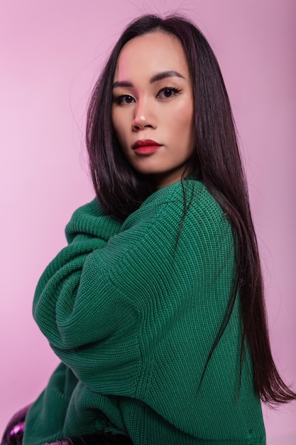 À la mode belle fille asiatique vietnamienne élégante avec des cheveux bruns et des lèvres rouges dans un chandail vert tricoté à la mode sur fond rose dans le studio Portrait féminin multicolore