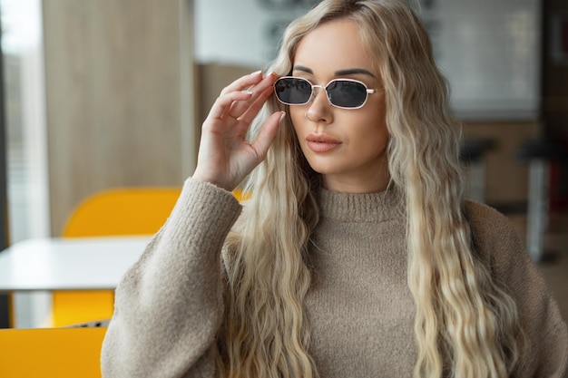 Mode belle fille blonde modèle dans un pull beige porte des lunettes de soleil élégantes vintage et est assis dans un café