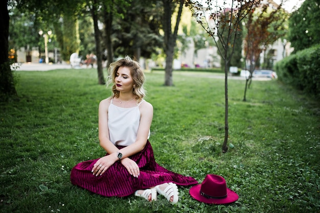 À La Mode Et Belle Fille Modèle Blonde En Velours Rouge élégant Jupe Chemisier Blanc Et Chapeau Assis Sur L'herbe Verte Au Parc