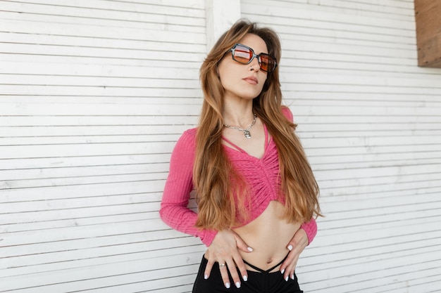 À la mode belle jeune femme élégante avec des lunettes de soleil colorées fraîches dans un haut court tricoté rose de mode avec une jupe noire se tient près d'un mur en bois blanc